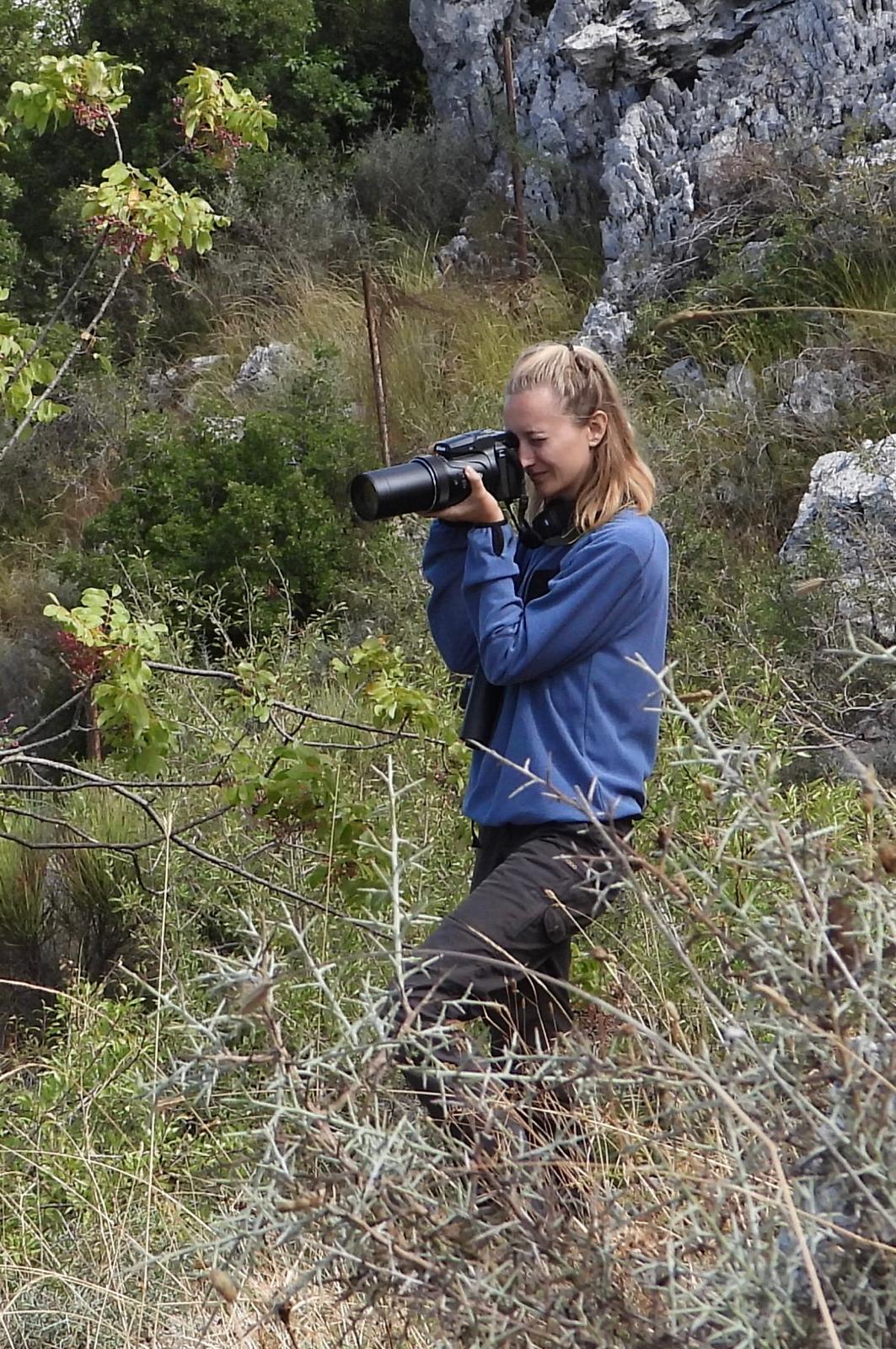 Alexandra holding a camera with a long lens among rocks and vegetation.