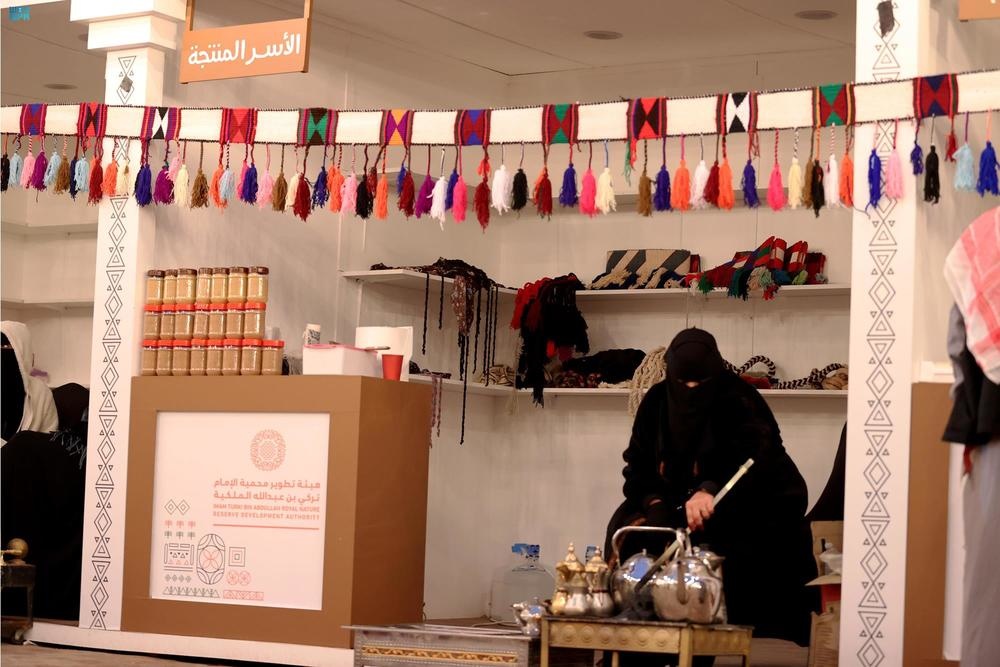 جناح للأسر المنتجة في شتاء درب زبيدة بلينة، تعرض فيه منتجات محلية ومنسوجات وحرف يدوية، مع سيدة تُعد القهوة. صورة أرشيفية منشورة في يناير 2022.
