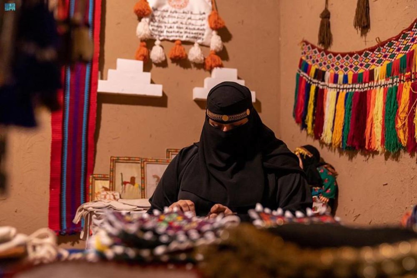 حرفية خلف طاولة مشغولات يدوية، تحيط بها منسوجات وزينة ملونة في شتاء درب زبيدة بمدينة لينة التاريخية. صورة أرشيفية منشورة في ديسمبر 2022.
